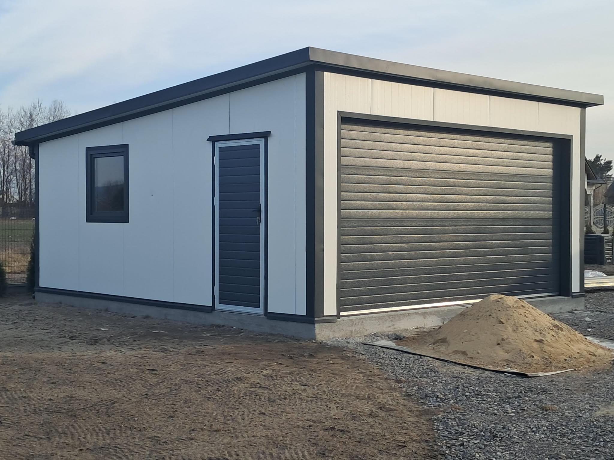 Biały garaż wolnostojący z ciemną bramą segmentową i drzwiami bocznymi